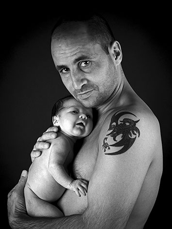 black and white portrait of a father holding shirtless and tattooed on the shoulder in her arms her baby son shot in studio Halet with a Mamiya 